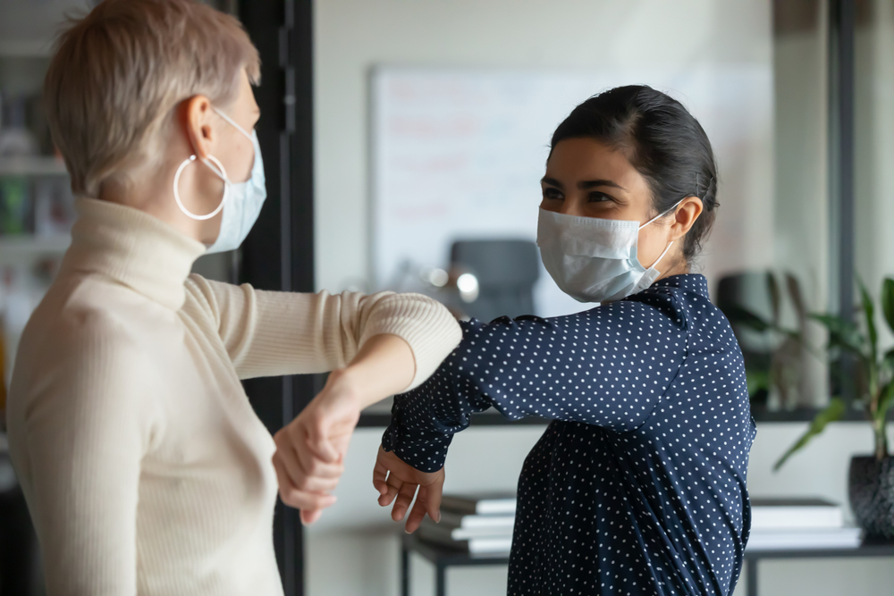 Smiling,Diverse,Female,Colleagues,Wearing,Protective,Face,Masks,Greeting,Bumping two women wearing masks elbow greeting